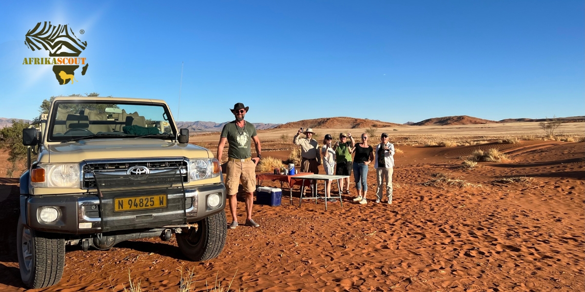 WiWi-SB-Lo Namibia, Namib-Wüste: Selbstfahrerreise mit 4x4-Geländewagen – Reisegruppe macht kurzen Stopp mit Tisch im roten Dünensand, weite Wüstenlandschaft und klarer blauer Himmel im Hintergrund.